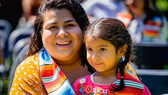 A graduating student who wears an orange shirt is smiling and holding her daughter in her lap while with her serape inspired graduate stole is wrapped around them both